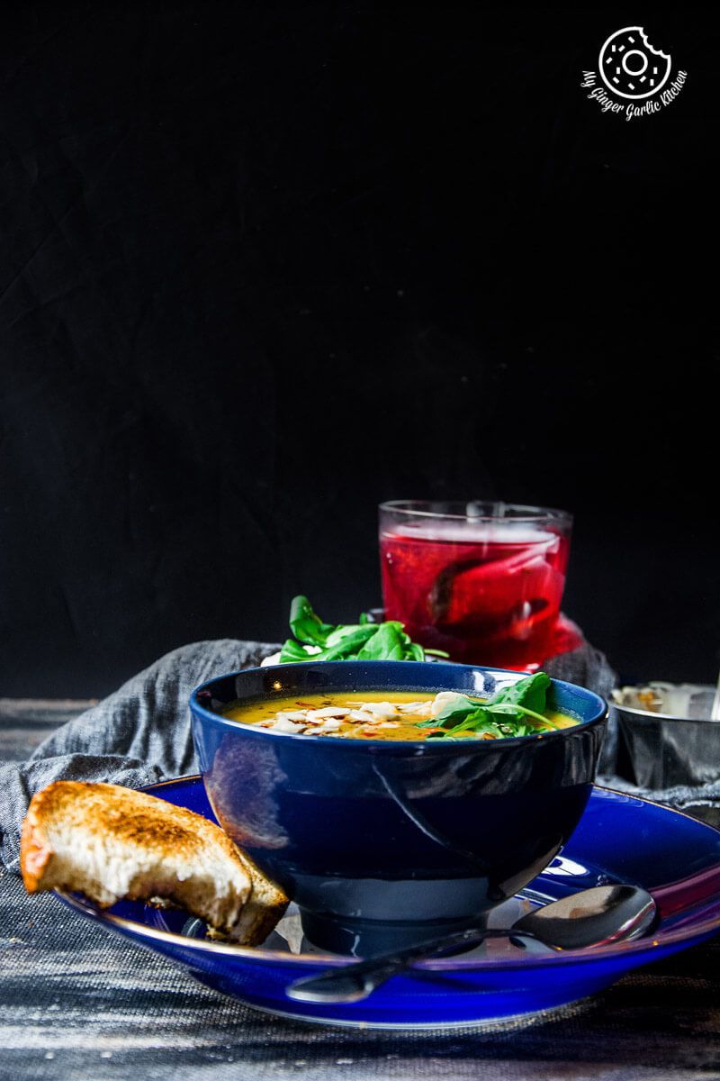 a blue bowl of sweet potato carrot soup with bread and a drink on the side