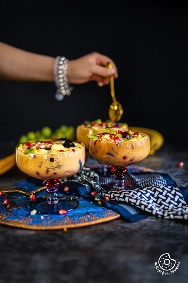 three fruit custard bowls in the frame and a hand holding a golden spoon over a fruit custard bowl