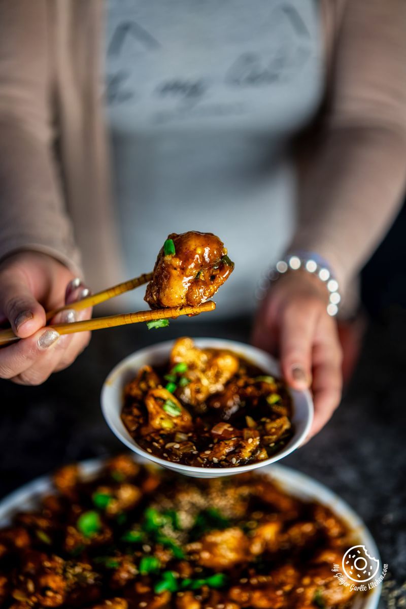 a female holding gobi manchurian with wooden chopsticks in one hand and a bowl with other hand