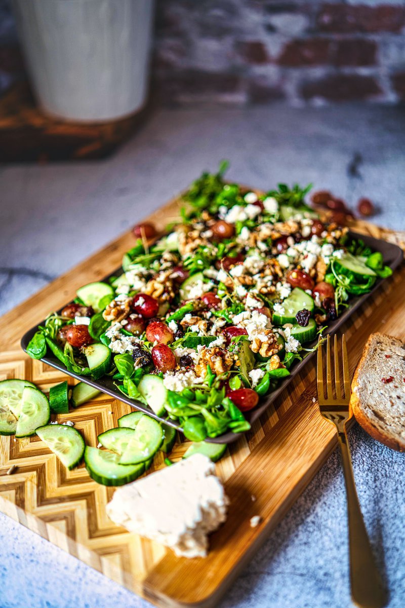 A vibrant grape salad with feta cheese and walnuts on a bamboo serving board, accompanied by sliced cucumber and a slice of multigrain bread.
