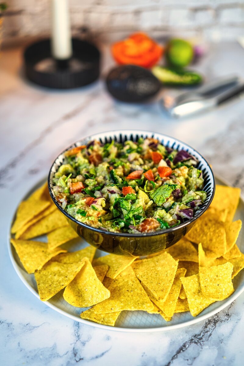 Freshly made chunky guacamole in a decorative blue and white bowl, surrounded by yellow corn tortilla chips on a white marble surface. Fresh ingredients visible in the background.