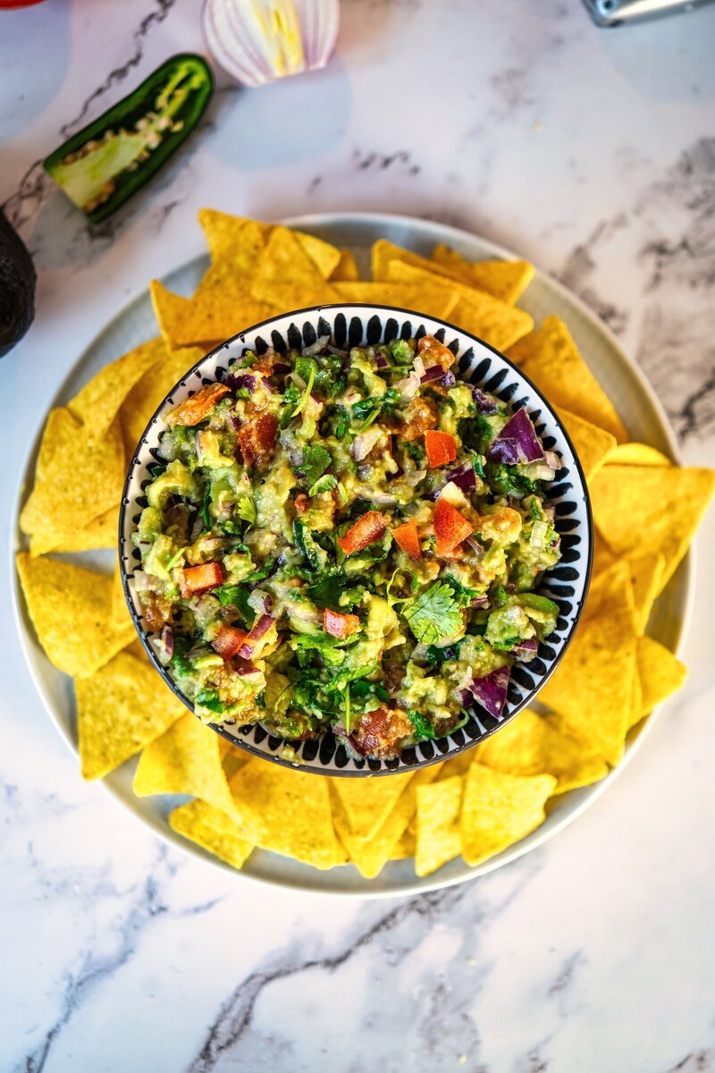 Overhead view of a bowl of guacamole with tomatoes and herbs, placed in the center of a plate surrounded by tortilla chips.