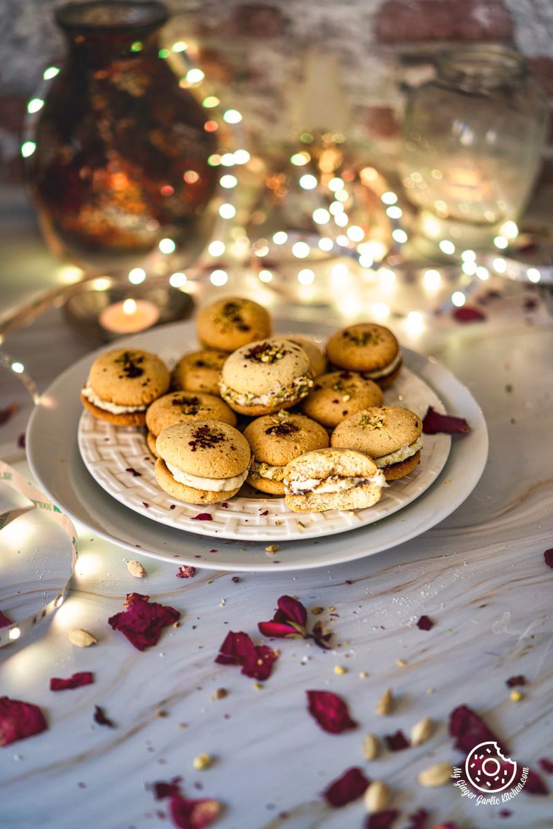 gulab jamun cookies in a white plate with some dried rose petals on the side and bouquet light in the background