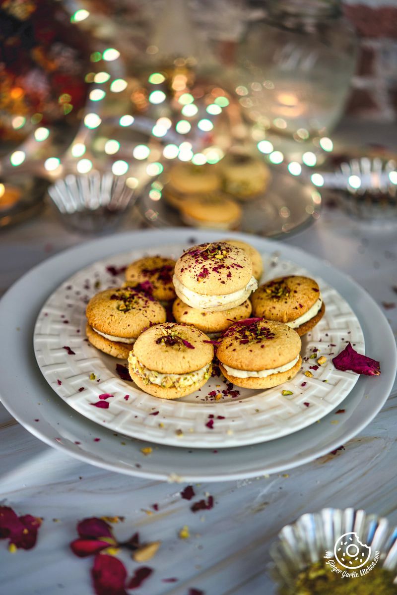 gulab jamun cookies stacked on a white plate with some dried rose petals on the side and bouquet light in the background