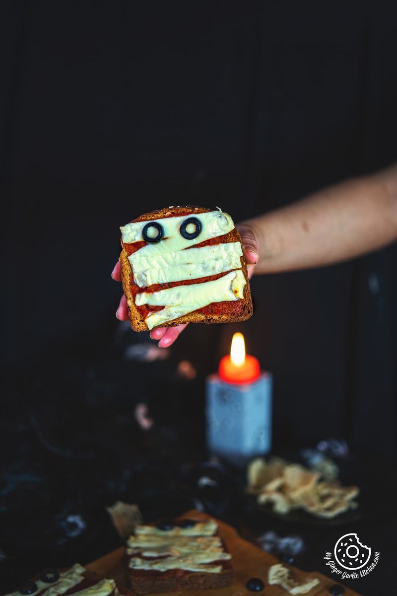 Close-up of a hand holding mummy pizza toast with a orange candle in background.
