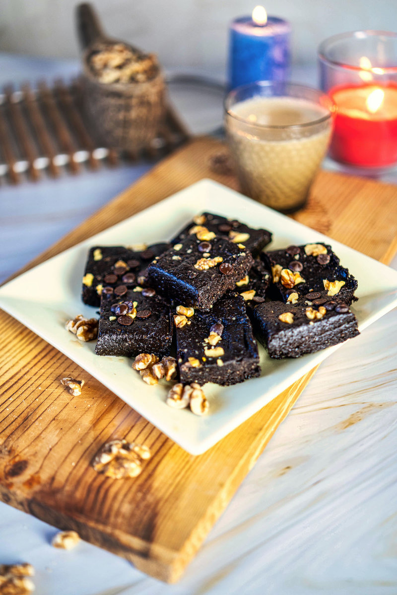 A plate of healthy date bars with nuts and chocolate chips on a wooden board, accompanied by a glass of milk and lit candles in the background.