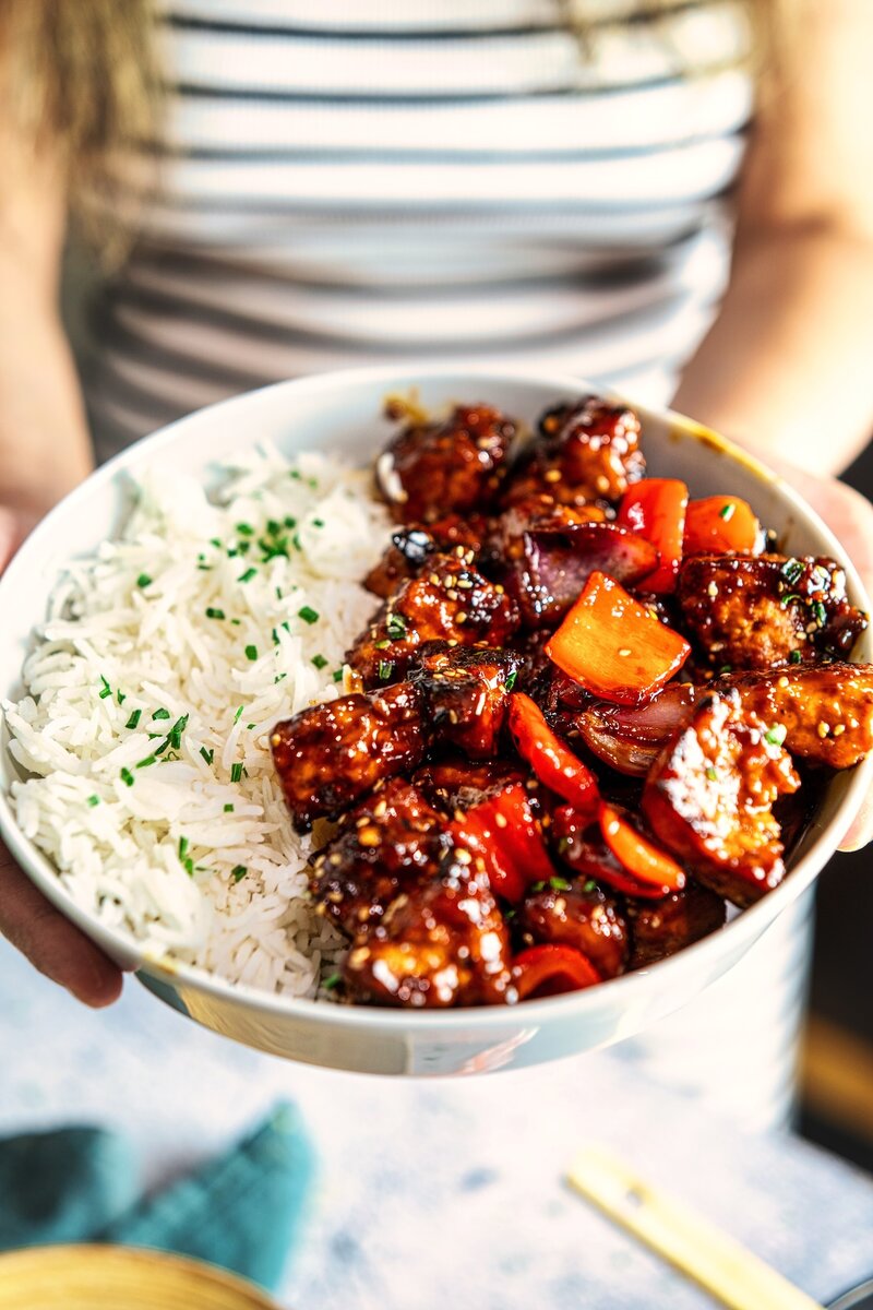 Bowl of crispy hoisin tofu served with steamed rice, garnished with fresh chives, held in hands