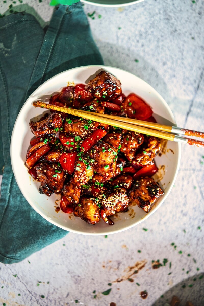 Crispy hoisin tofu stir fry with red bell peppers, sesame seeds, and chives in a white bowl with chopsticks