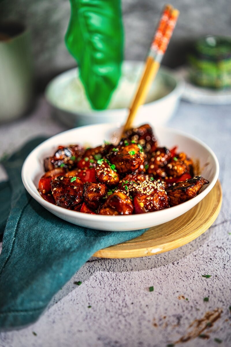 Side view of crispy hoisin tofu stir fry served in a white bowl over a teal napkin, topped with sesame seeds and chives