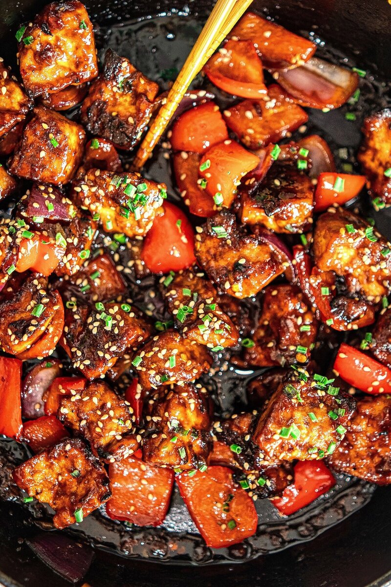 Close-up of glossy crispy hoisin tofu with red peppers and onions in a cast iron pan, sprinkled with sesame seeds and chives