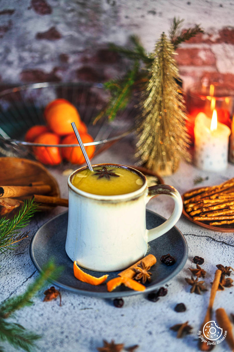 there is a cup of hot orange juice with cinnamon, oranges, candles and christmas tree in the background