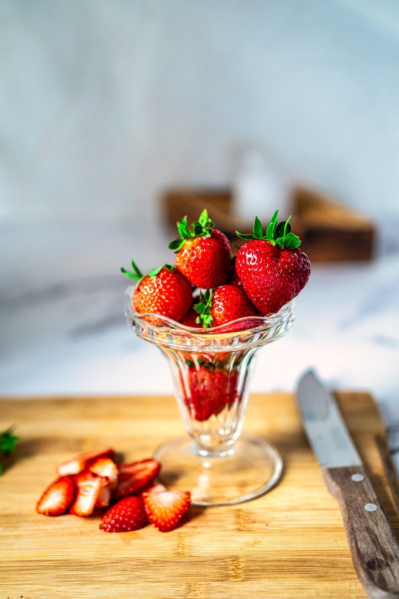 A glass serving bowl filled with fresh, whole strawberries on a wooden cutting board, with a kitchen knife and sliced strawberries visible in the background