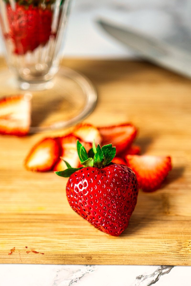 Close-up of a single ripe strawberry on a wooden cutting board, with sliced strawberries and a glass serving bowl in the background.