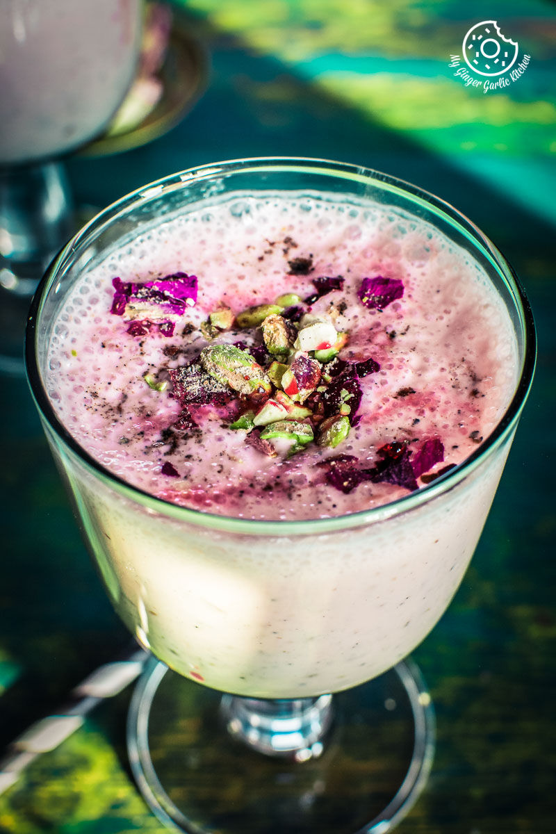 a glass of rose lassi topped with rose syrup and chopped nuts