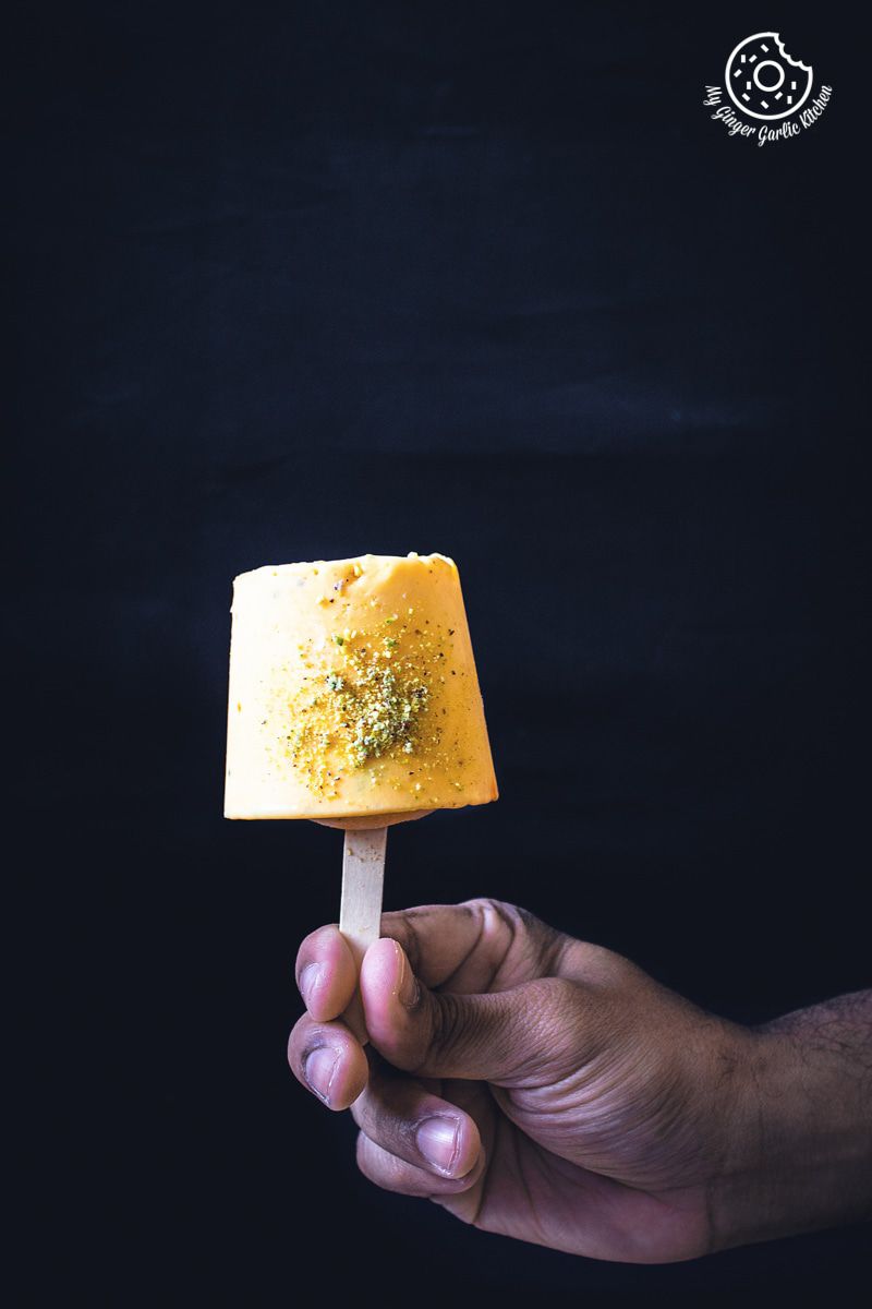 someone holding a mango kulfi popsicle with a pistachio topping on it