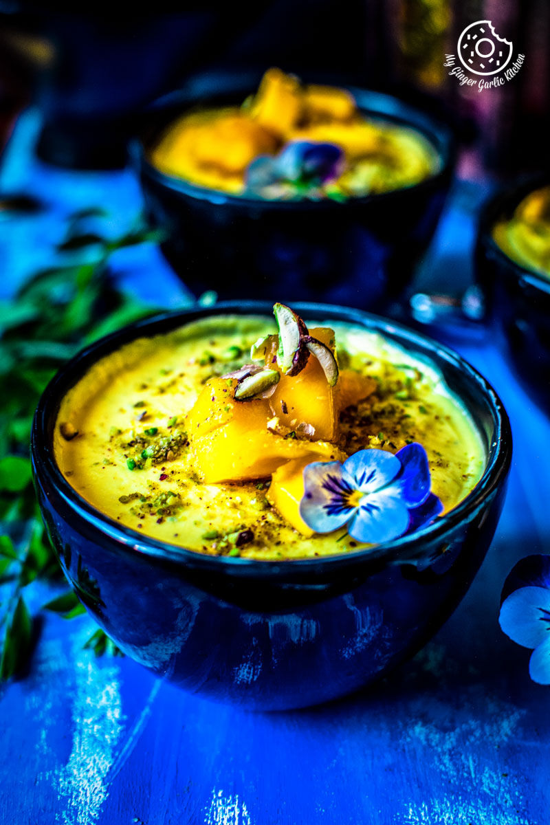 bowls of mango shrikhand with a blue flower, on a blue background and some leaves on the side