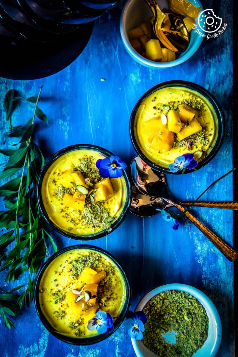 three bowls of mango shrikhand topped with mangoes, flowers, pistachios with spoons