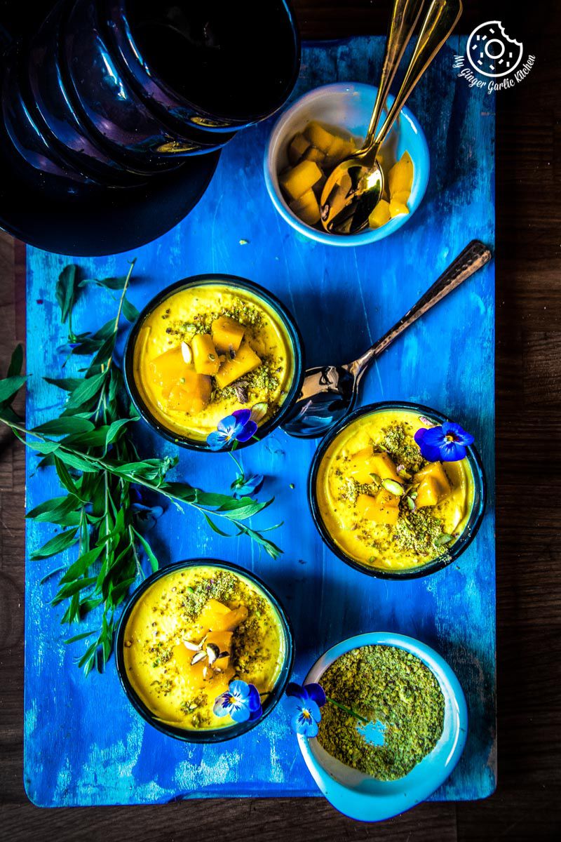 three bowls of mango shrikhand topped with mangoes, flowers, pistachios with spoons on a blue tray with spoons