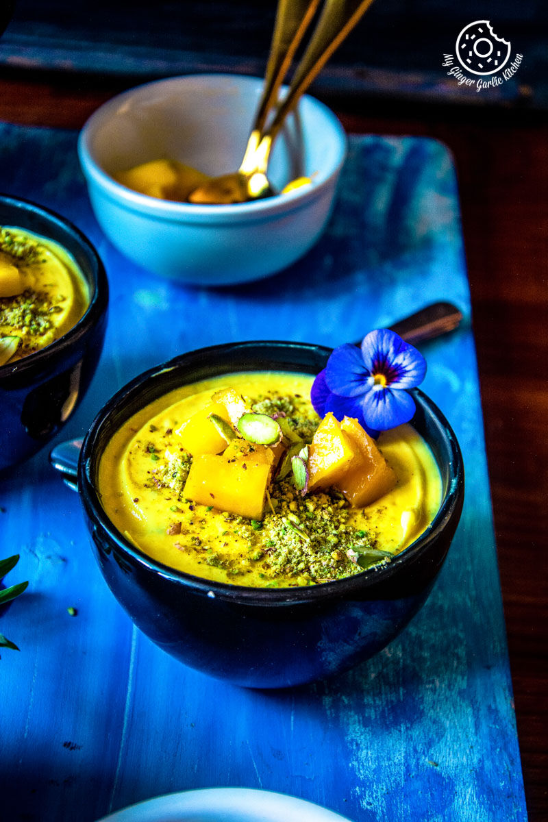 a bowl of mango shrikhand topped with mangoes, flowers, pistachios on a blue table with a flower in it