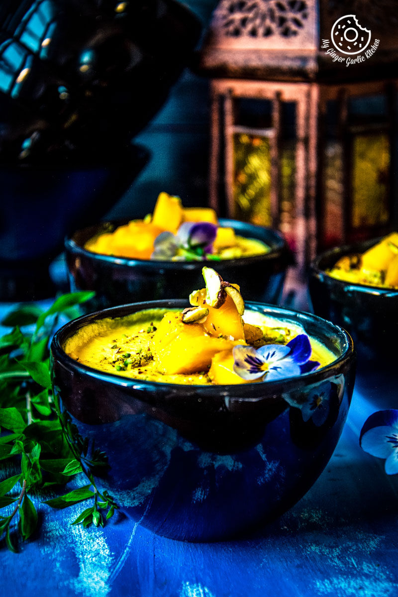 two bowls of garnished mango shrikhand on a blue table with a candle