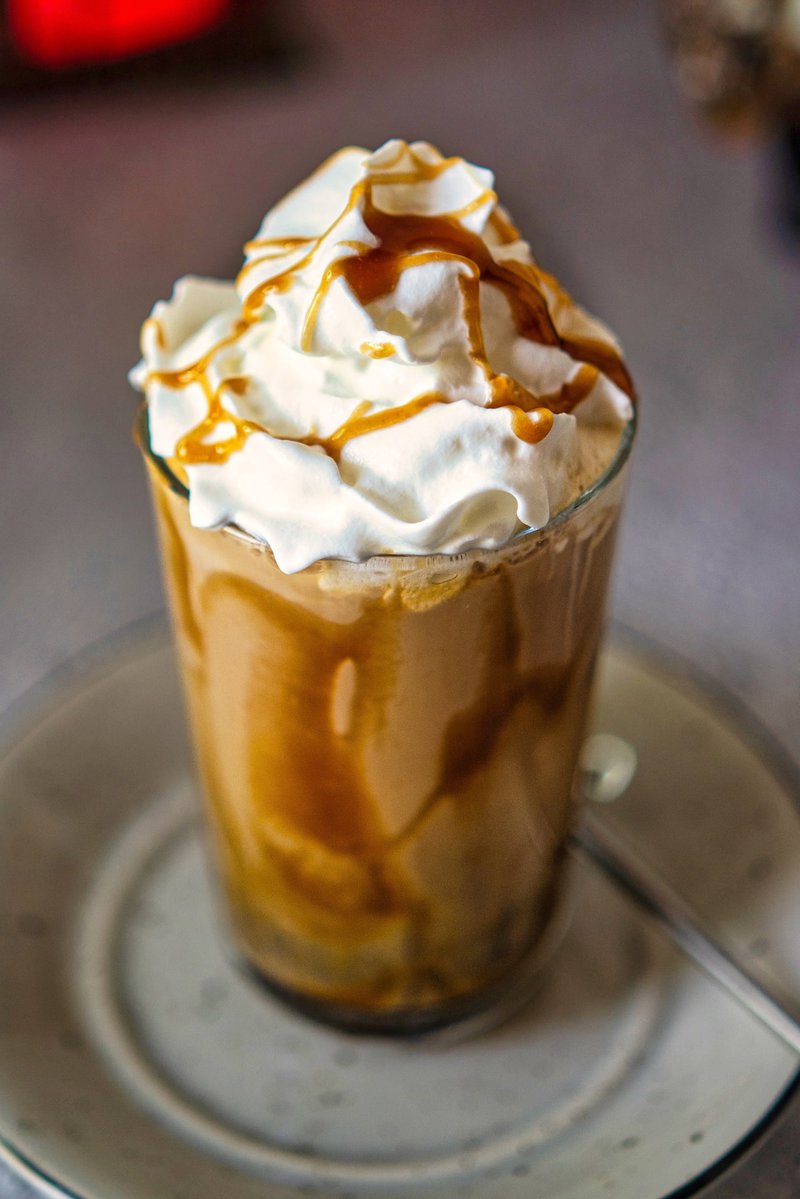 Iced caramel latte topped with a generous amount of whipped cream and caramel sauce, served in a glass on a saucer.