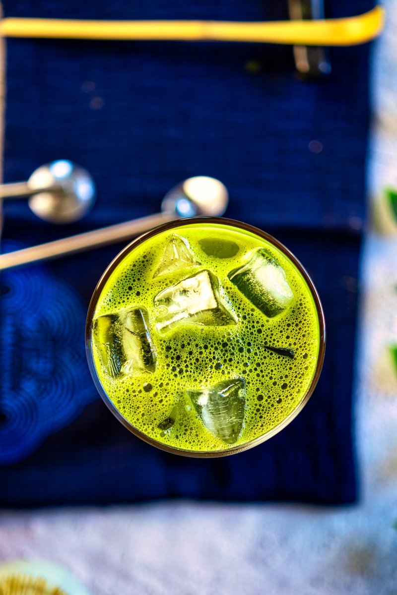 Top view of a frothy iced matcha latte with ice cubes served on a dark blue cloth with matcha tools