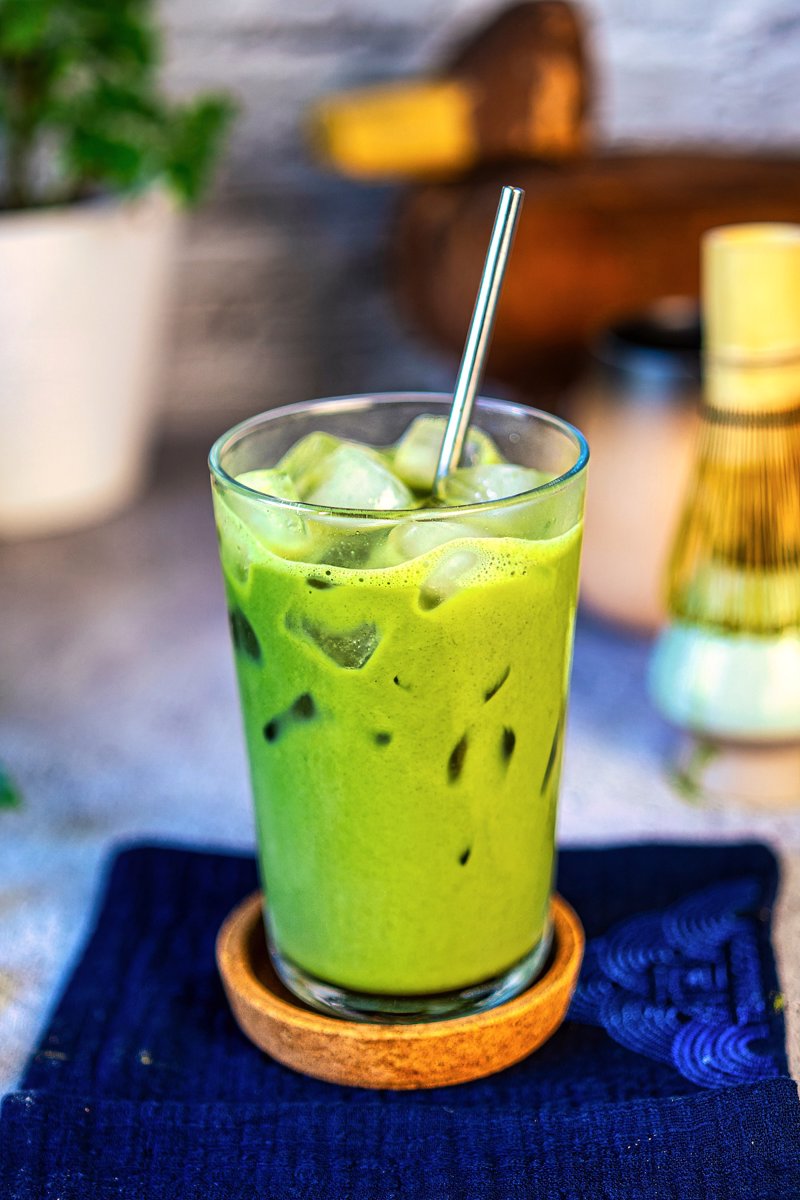 Refreshing iced matcha latte with ice and a straw served with a bamboo matcha whisk in the background