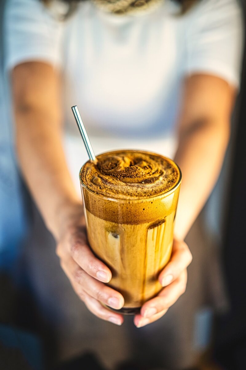 Hands holding iced oat milk dalgona latte with thick whipped coffee foam and layered texture