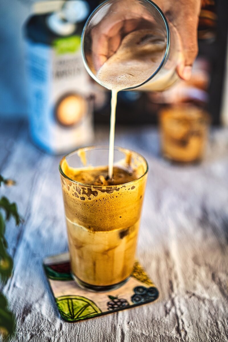 Pouring oat milk into iced dalgona coffee creating layered vegan iced latte with creamy foam