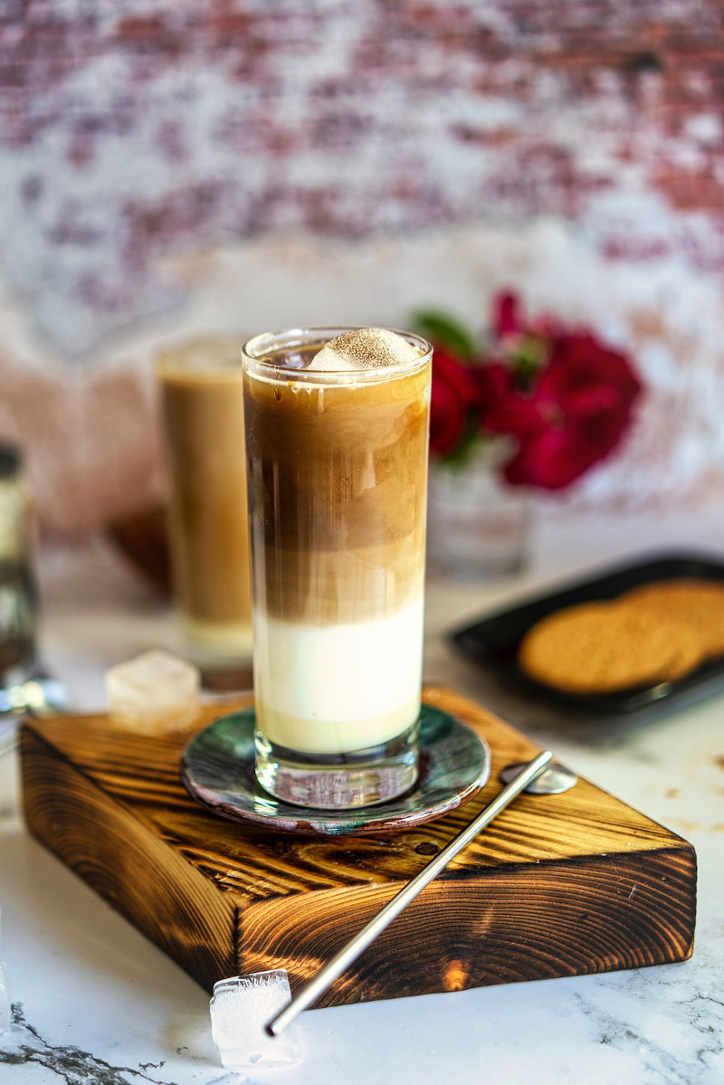 A beautifully layered Iced Spanish Latte in a tall glass, resting on a wooden coaster, with a rustic background.