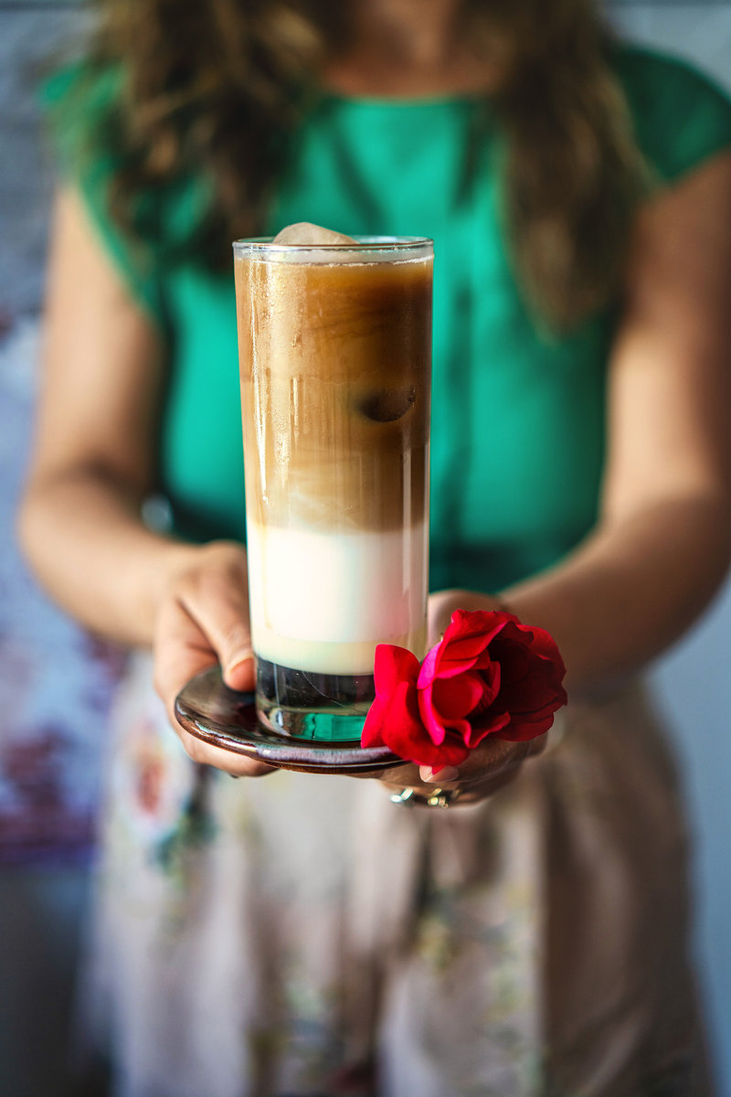 A person holding a tall, layered Iced Spanish Latte with a red flower, highlighting the creamy texture and aesthetic presentation.