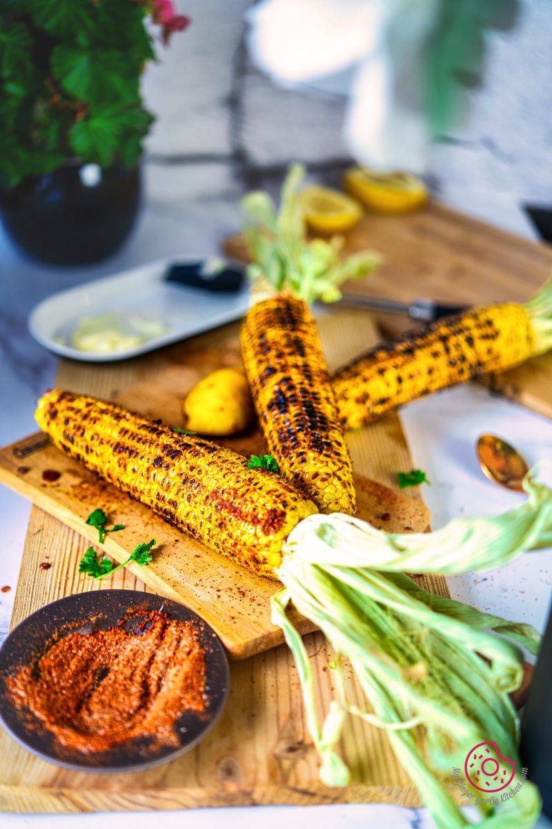 3 indian roasted corn on the cobs or bhutta masala on a wooden board with a bowl of spice