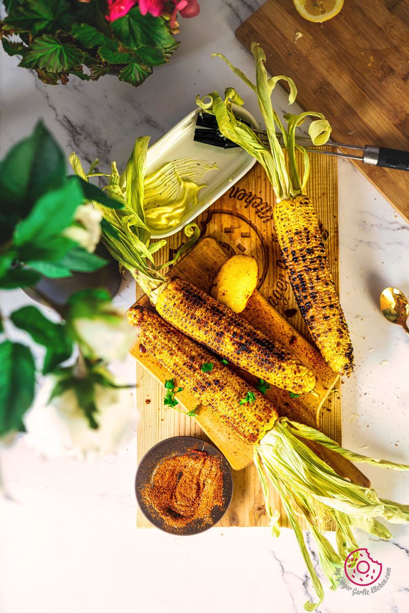 flatlay of 3 indian style roasted corn on the cobs or masala bhutta on a wooden board with a bowl of spice