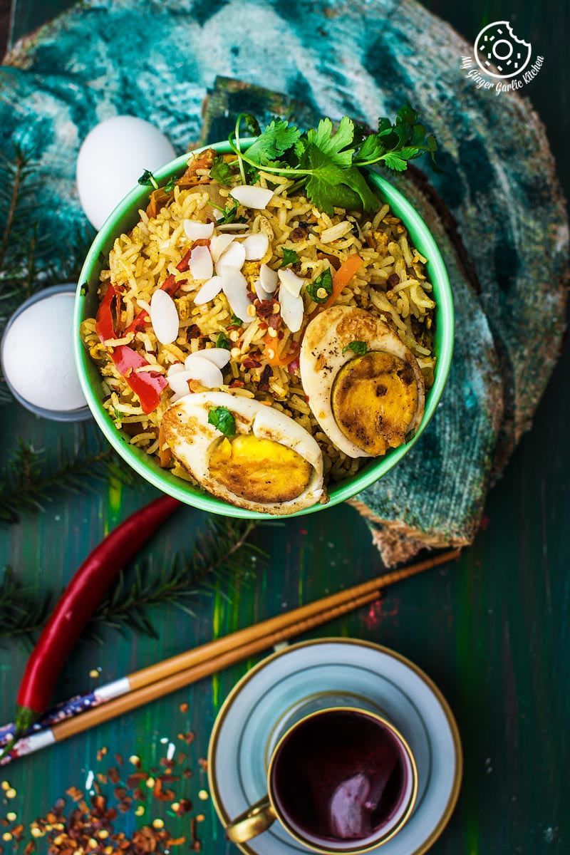 a bowl of indian style triple egg fried rice with almond flakes, chopsticks and a cup of tea, and eggs on the side