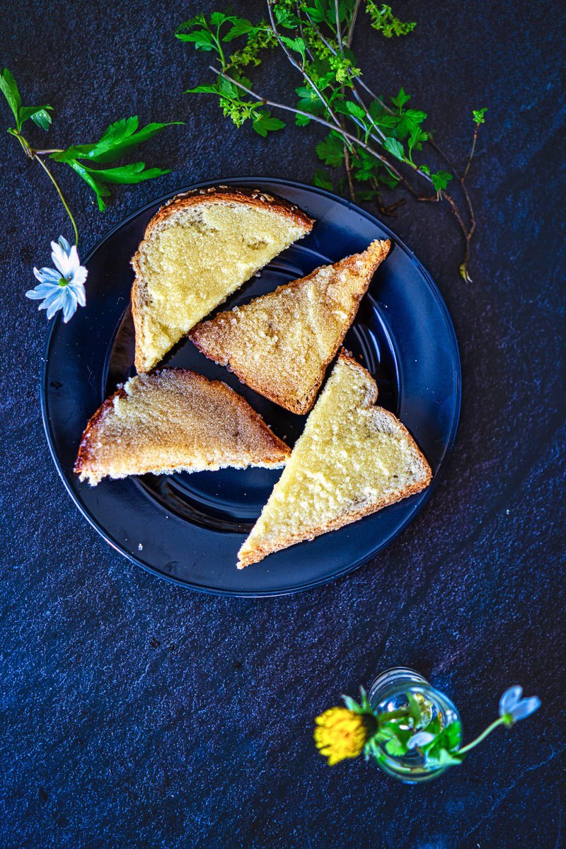 Indian sweet bread garnished with sugar on a dark plate with fresh green herbs and white flowers on a dark textured surface.