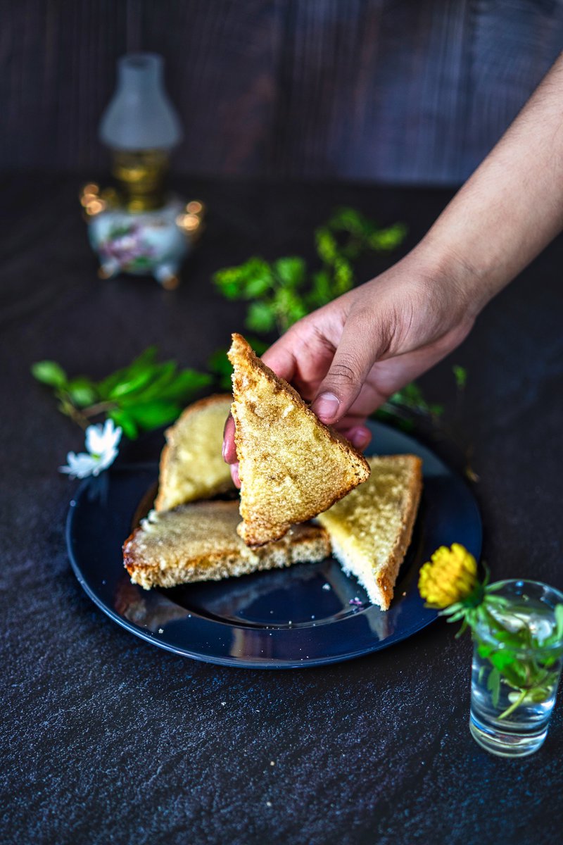 A hand picking up a slice of Indian sweet bread from a dark plate, with herbs and a small lamp in the background.