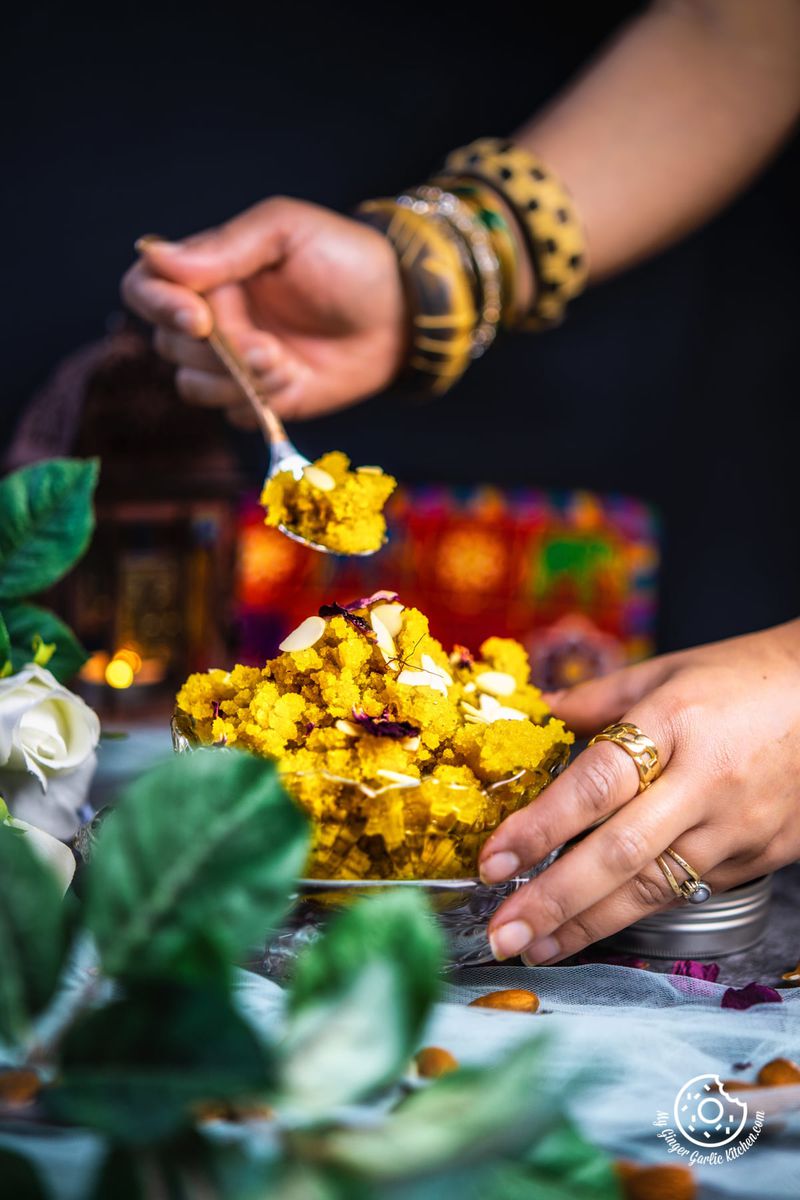 a female holding a spoon filled with badam halwa over instant badam halwa transparent bowl