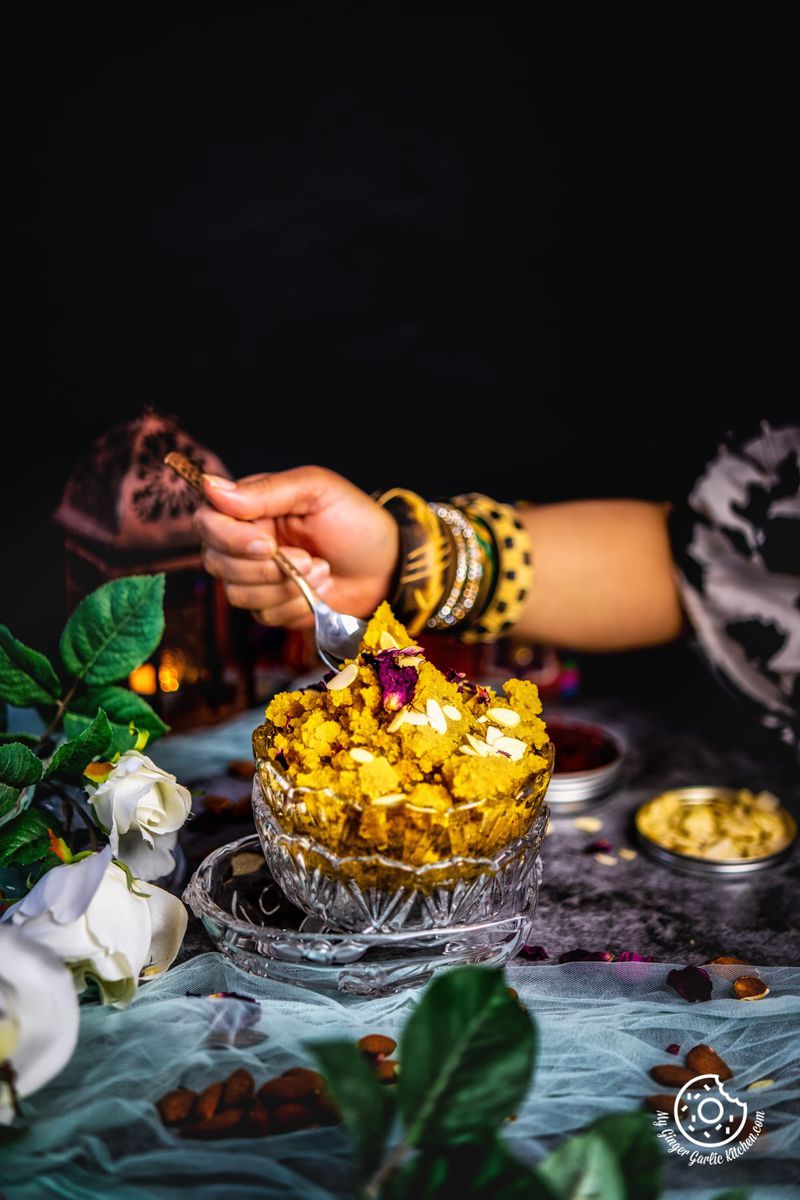 a feamle holding a spoon filled with badam halwa over instant badam halwa transparent bowl