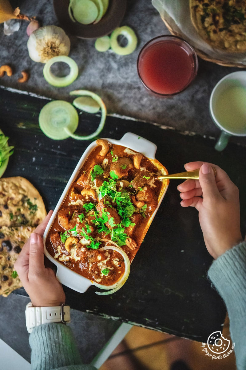 overhead shot of a female holding zucchini tofu curry served in a white rectangular casserole dish from one hand and other hand holding a golden spoon