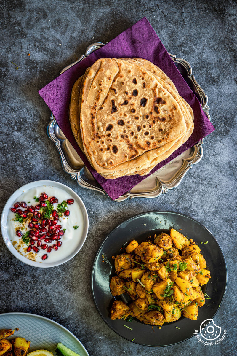 overhead shot of jeera aloo served in a grey plate with  pomegranate raita and parathas on the side