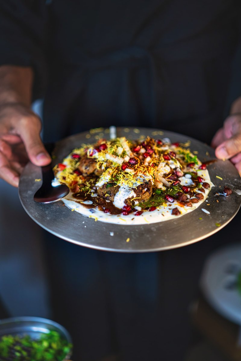 Person holding a plate of Kala Chana Tikki Chaat garnished with yogurt, sev, and pomegranate seeds.