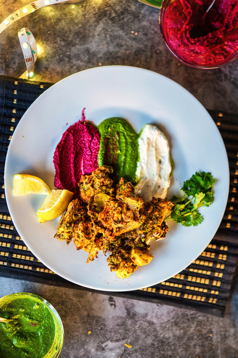 A plate of kale pakoras served with vibrant beetroot, green chutney, white dip, lemon wedges, and cilantro garnish.