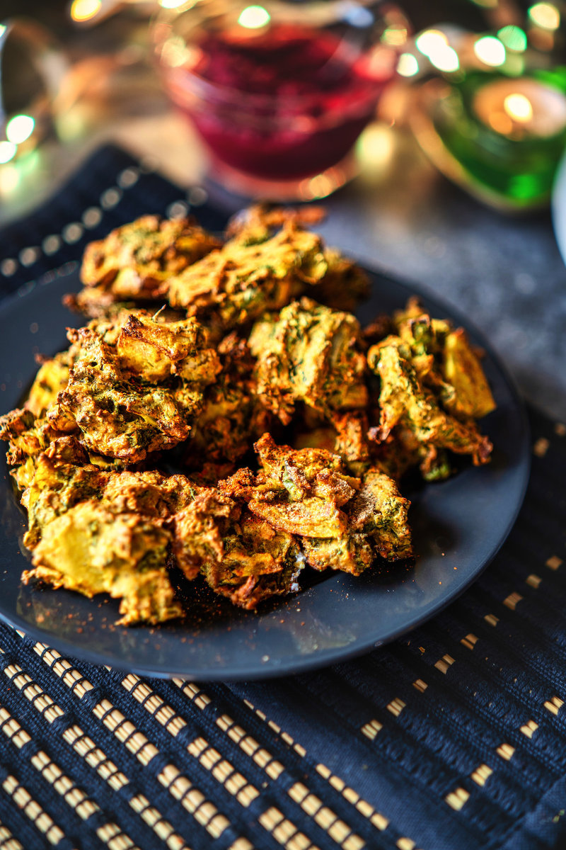 A plate of crispy kale pakoras on a patterned tablecloth, with colorful glasses and lights in the background.