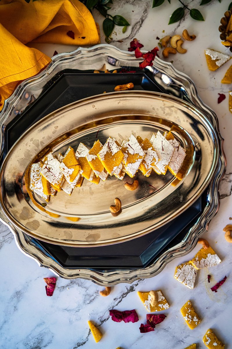 Silver platter with diamond-shaped saffron kaju katli sweets arranged in a line, surrounded by nuts and flower petals.