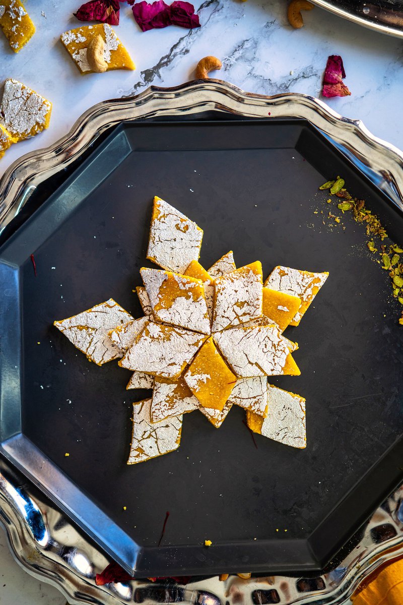 Diamond-shaped kesar kaju katli sweets on a dark grey tray, with flower petals and nuts scattered around.