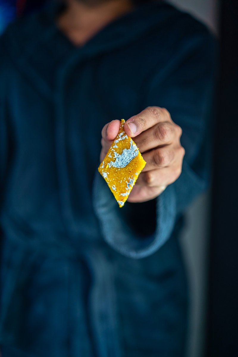 Hand holding a diamond-shaped kesar kaju katli covered in silver foil against a dark blue background.