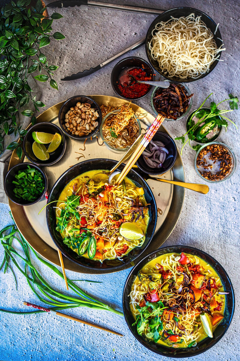 Traditional Burmese Khow Suey recipe with coconut curry noodles and toppings, served with assorted condiments and garnishes on a tabletop