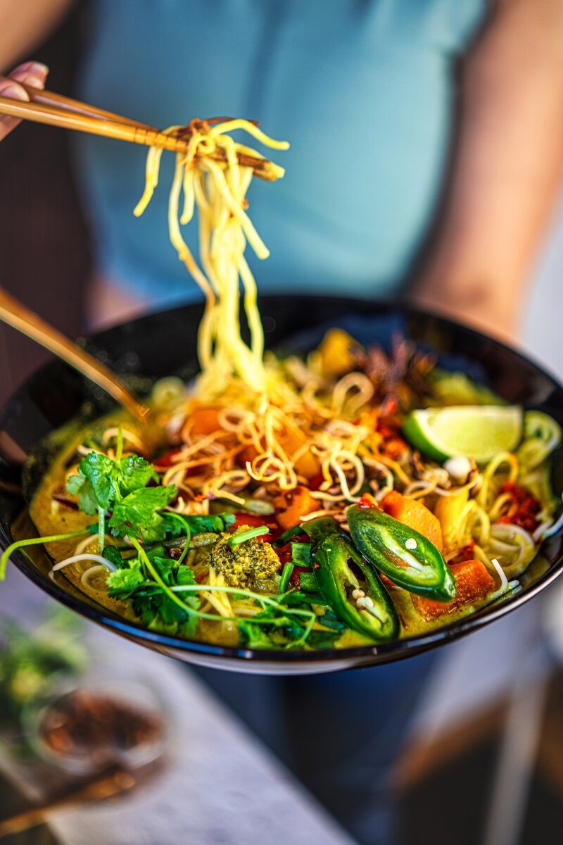 Steaming hot Khow Suey noodle curry being served with chopsticks featuring yellow egg noodles, coconut broth, green herbs, and jalapeños in authentic Burmese street food style