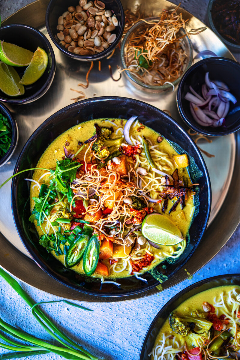 Authentic Burmese Khow Suey curry noodle soup with crispy noodles, vibrant vegetables, fresh lime, and traditional garnishes in a black bowl served with condiments for customizing flavor