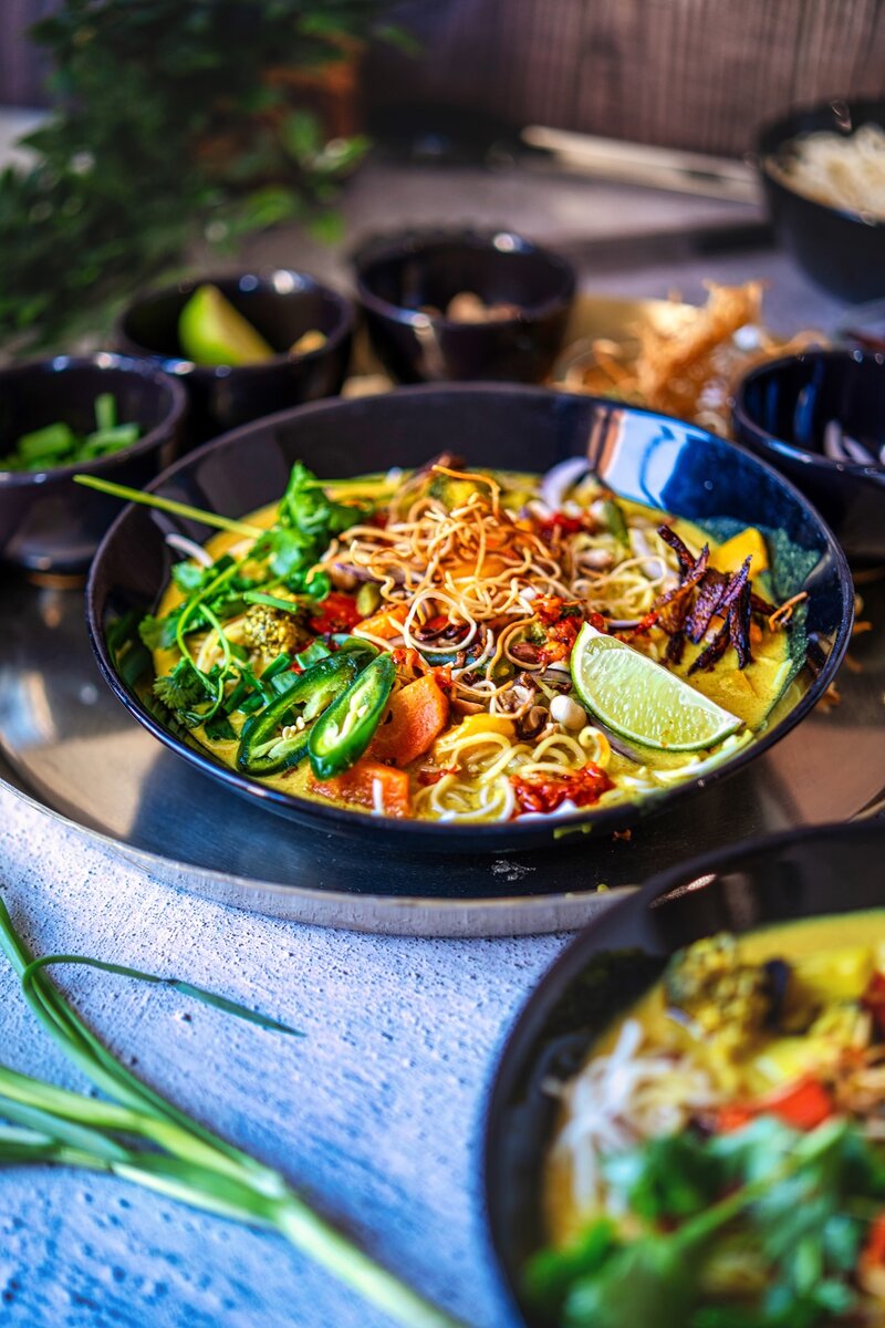 IHomemade Khow Suey noodle soup in a black bowl with coconut curry, crispy noodles, lime wedges, onions, green chilies, and fresh herbs